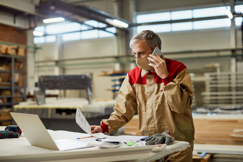Ouvrier en tenue professionnelle utilisant un téléphone et un ordinateur portable dans un atelier industriel.