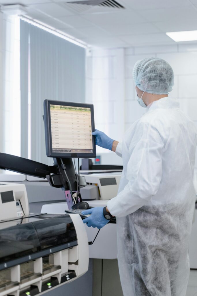 Technicien en blouse et charlotte travaillant sur une machine d'analyse automatisée et consultant un écran d'ordinateur dans un environnement contrôlé, illustrant le besoin d'équipements électroniques certifiés.